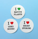 Three round white stickers on a light blue background. Top Sticker reads: I LOVE NATIVE PLANTS with love being represented by a green heart. Beneath reads I LOVE USED BOOKS with love being represented by a red heart. To the right reads I LOVE SHORT STORIES with love being represented by a red heart.