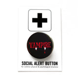 A round black pinback button that reads VAMPIRE in a spooky red font with white fangs dripping with blood peeking out at the bottom of the word.  Button is on a Social Alert Button backing card.