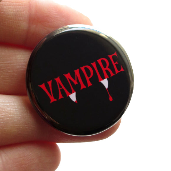 A round black pinback button that reads VAMPIRE in a spooky red font with white fangs dripping with blood peeking out at the bottom of the word.  Button is held in a hand.