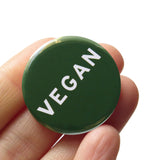 A round dark green (the color of leafy Dino kale) pinback button that reads VEGAN in white text. Button is held in a hand.