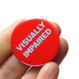 a round red 1.25" pinback button reads: VISUALLY IMPAIRED in light blue text. Button is held in a hand.