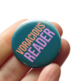 Round teal colored pinback button which reads Voracious Reader in sans serif font. Voracious is in peach, Reader is a larger word and in lavender. The button is held in a hand.