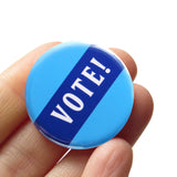 lue round pinback button that reads VOTE! in white across a navy blue diagonal strip. Button is held in a hand.