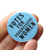 Light blue round pinback button that reads VOTES FOR PROGRESSIVE WOMEN! in black text. Button is held in a hand.