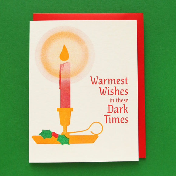 A 3-color risograph holiday card that features a red candle with a round yellow glow, dripping wax, in a classic gold holder with a spring of green holly berries. The red text reads Warmest Wishes in these Dark Times. A red envelope is with the card, and they sit on a green background.