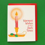 A 3-color risograph holiday card that features a red candle with a round yellow glow, dripping wax, in a classic gold holder with a spring of green holly berries. The red text reads Warmest Wishes in these Dark Times. A red envelope is with the card, and they sit on a green background.
