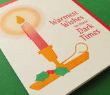 Angled close up of a 3-color risograph holiday card that features a red candle with a round yellow glow, dripping wax, in a classic gold holder with a spring of green holly berries. The red text reads Warmest Wishes in these Dark Times. A red envelope is with the card, and they sit on a green background.