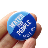 A round blue 1.25" pinback button that reads WATER FOR PEOPLE, NOT AIin 2 different white fonts. Badge is held in a hand. 
