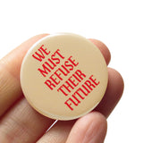A round cream colored pinback button that reads WE MUST REFUSE THEIR FUTURE in red-orange text. Badge is held in a hand.