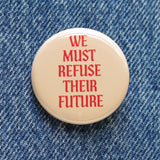 A round cream colored pinback button that reads WE MUST REFUSE THEIR FUTURE in red-orange text. Button is pinned to blue denim.