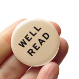 Round 1.25" cream colored pinback button that says WELL READ in black text. Badge is held in a hand.