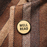 Round enamel pin that reads WELL READ in two lines in sans serif text. Gunmetal silver text and outline, on a cream enamel background. Pin is on the lapel of a brown tweed blazer.