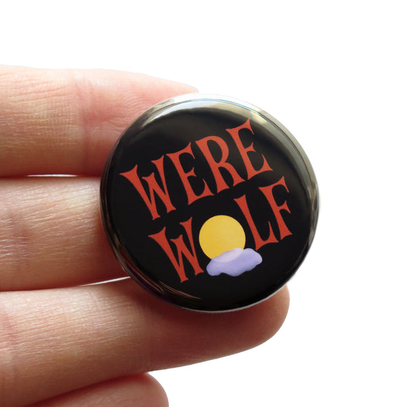 A round black pinback button that reads WEREWOLF in a spooky brown text with the O as a moon with a foggy cloud behind it. .Button is held in a hand.