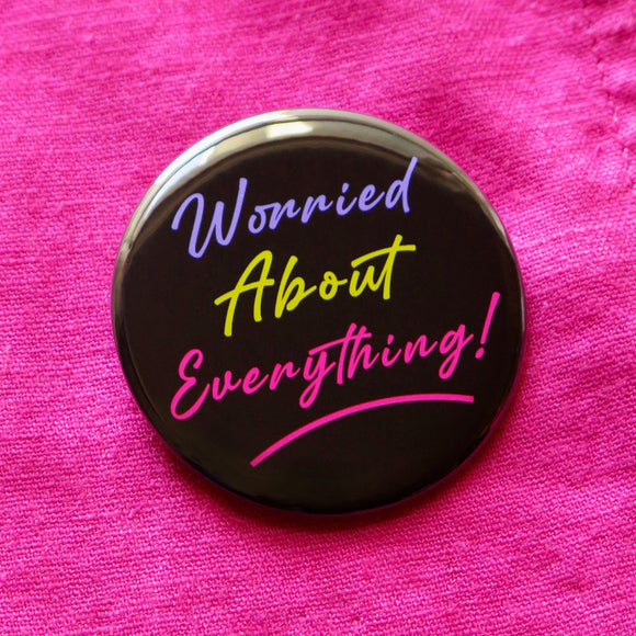 A round black  pinback button that reads WORRIED ABOUT EVERYTHING!  in purple, neon green & magneta pink text, in a retro 80's vibe.  Button is pinned to bold pink linen fabric.