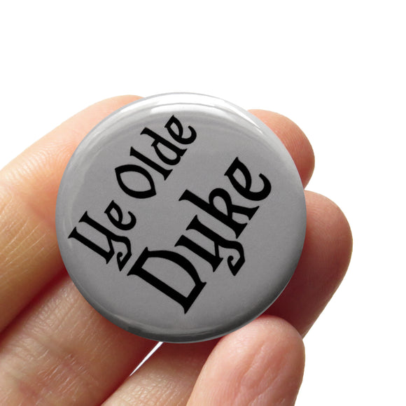 Round gray pinback button that reads YE OLD DYKE in a black old English front. Badge is held in a hand.