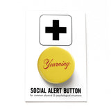 A round butter yellow 1.25" pinback button that reads " Yearning" in a Burgundy script font. The button is on a white card with a black plus sign and the text 'SOCIAL ALERT BUTTON for common physical & psychological situations'.