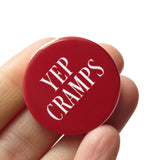 A round burgundy pinback button that reads YEP CRAMPS in think white text. Button is held in a hand.