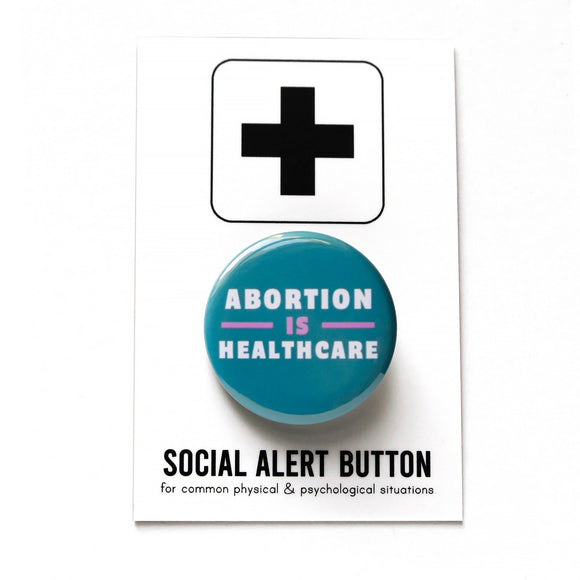 A round teal-blue pinback button that reads ABORTION IS HEALTHCARE in three lines in white & lavender text. Badge is pinned to a Social Alert Button backing card, with a plus sign at the top.