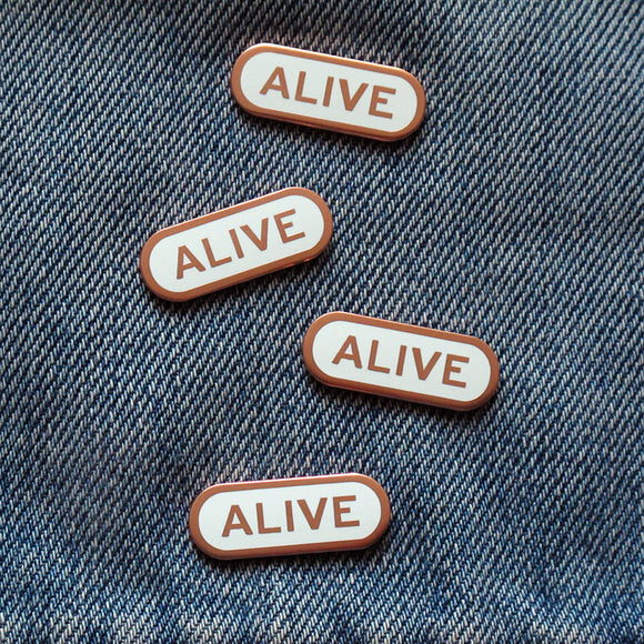 Four pill shaped white enamel pins with a rose gold copper outline and rose gold text reading ALIVE, cascading down a piece of blue denim.