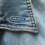 Capsule shaped enamel pin that says BIBLIOPHILE on the lapel of a blue denim jacket. 