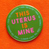 Big 3 inch shiny olive green pinback button with bright orange and hot pink chunky san serif text that reads: This Uterus is Mine. Smaller text in a light blue runs in a circle around the permitter that says Fuck You Alito, Fuck You Thomas, Fuck you Coney-Barrett, Fuck you Gorsuch, Fuck you Kavanaugh. The button is pinned to a bright orange sweater shirt. 