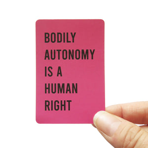 A vertical upright sticker with rounded corners, reads BODILY AUTONOMY IS A HUMAN RIGHT in black san serif text on a mauve-raspberry background