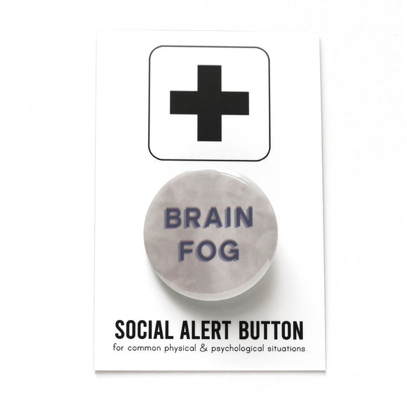 Round pinback button that says BRAIN FOG in blue-gray sans serif text on a cloudy gray background. Button is a on a white backing card that reads SOCIAL ALERT BUTTON on the bottom, with a black plus sign at the top.
