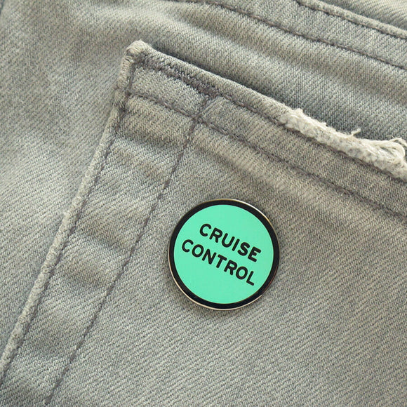 Round enamel pin that says CRUISE CONTROL on a grey denim pocket.
