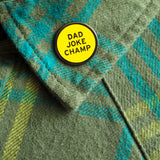 Round enamel pin the says DAD JOKE CHAMP on the lapel of a forrest green plaid flannel shirt