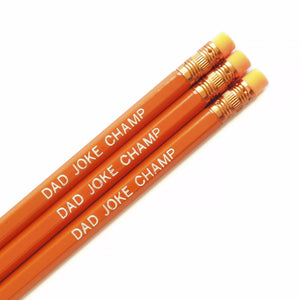 A white square, with the top half of three orange pencils with orange ferrules and orange erasers, in a diagonal position. Stamped with the phrase: DAD JOKE CHAMP in silver foil.