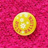 Round, lime green pinback button that reads DILLY DALLIER in deep pink, in a half circle around the edge. The rest of the button has little white daisies with yellow centers.  Button is on a fascia sweater.