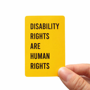 A golden yellow, vertical rectangle sticker with rounded corners is held in the lower right hand corner with a thumb and forefinger. The stickers reads DISABILITY RIGHTS ARE HUMAN RIGHTS in a black san serif text