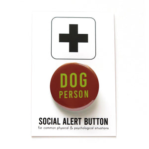 Round reddish-brown pinback button that reads DOG PERSON in lime green san serif text. Button is on a Social Alert Backing Card with a black plus sign at the top.