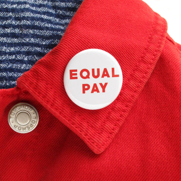 Round white pinback button that reads EQUAL PAY in red text. Badge is the lapel of a red jean jacket.