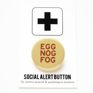Product shot of a 1.25" holiday themed pinback button that says EGG NOG FOG in brown text on a cream colored background. Button is on a Social Alert Button backing card.