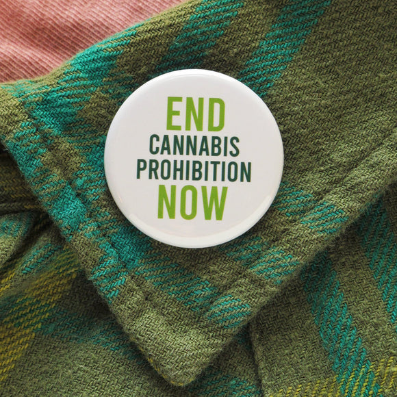 A round white pinback button that reads End Cannabis Prohibition Now, in light & dark green text.  Badge is pinned to a green plaid flannel shirt lapel.