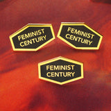 Three hexagon shaped hard enamel lapel pins that say FEMINIST CENTURY on a mottled red background.