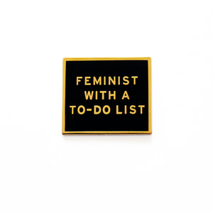 Rectangle enamel pin that says FEMINIST WITH A TO-DO LIST.  Gold text and outline on a black enamel background