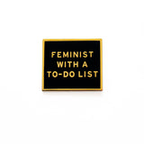 Rectangle enamel pin that says FEMINIST WITH A TO-DO LIST.  Gold text and outline on a black enamel background