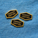 Three hexagon shaped hard enamel lapel pins that say FEMINIST CENTURY on a blue denim background.