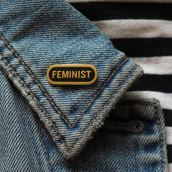 Capsule shaped enamel pin that says FEMINIST on the lapel of a blue denim jacket.