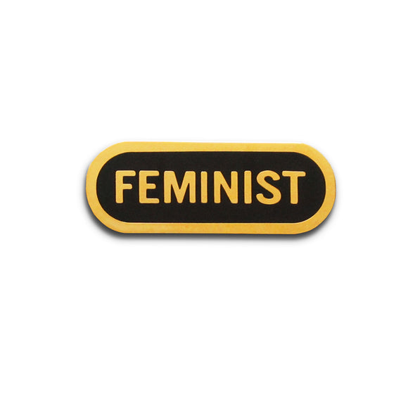Capsule shaped enamel pin that says FEMINIST.  Gold text and outline on a black enamel background