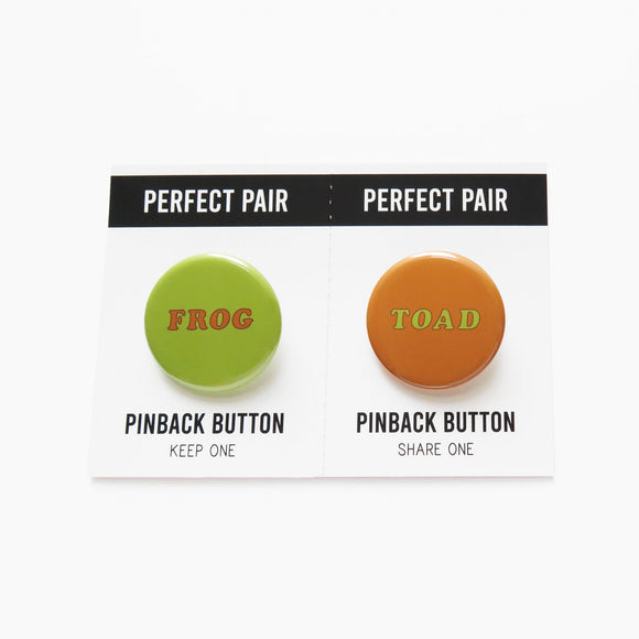 Two pinback buttons on a backing card with a center perforation to tear & share with a friend. Button on the left reads FROG with a light green background and brown text. Right sided button reads TOAD in light green with a brown background.