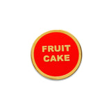 small circular enamel pin, bright red enamel background with gold text that reads FRUIT CAKE across two lines. Gold colored outline.
