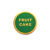 small circular enamel pin, green enamel background with gold text that reads FRUIT CAKE across two lines. Gold colored outline.