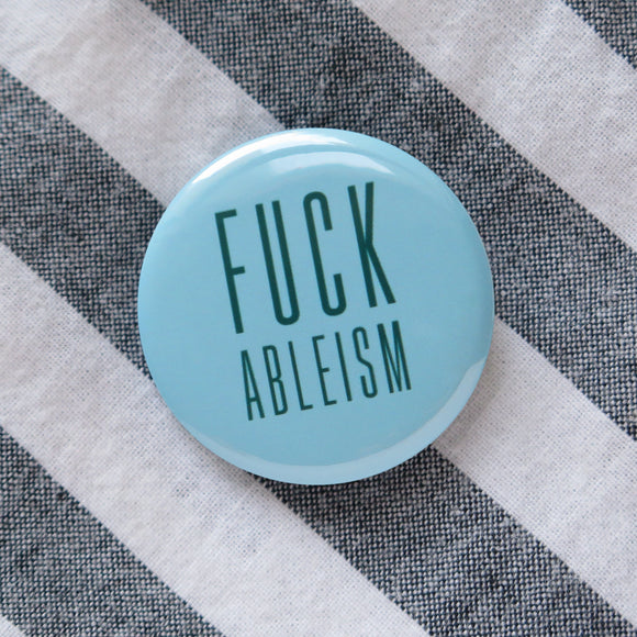 A round pale teal pinback button that reads FUCK ABLEISM in a dark teal color, in a condensed sans serif font.  Button is pinned to a gray & white diagonal stripped cotton shirt.