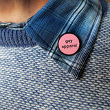 Small circular pink pin that reads Gay Apparel. Pin is pinned on a blue patterned shirt lapel.