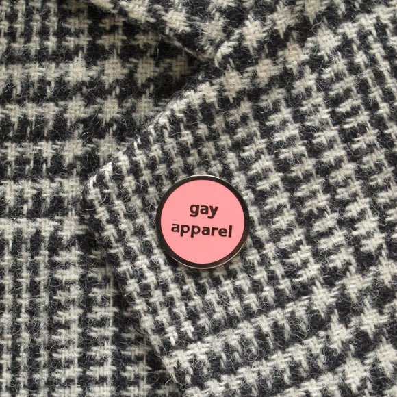 Small circular pink enamel pin with gunmetal silver text and outline that reads GAY APPAREL. Pin is pinned on a black and white checked coat.