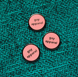 Three small pale pink enamel pins that read GAY APPAREL in gunmetal silver. Pins are pinned on a teal & black patterned fabric.