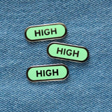 Three capsule shaped enamel pins that say HIGH on a blue denim background.